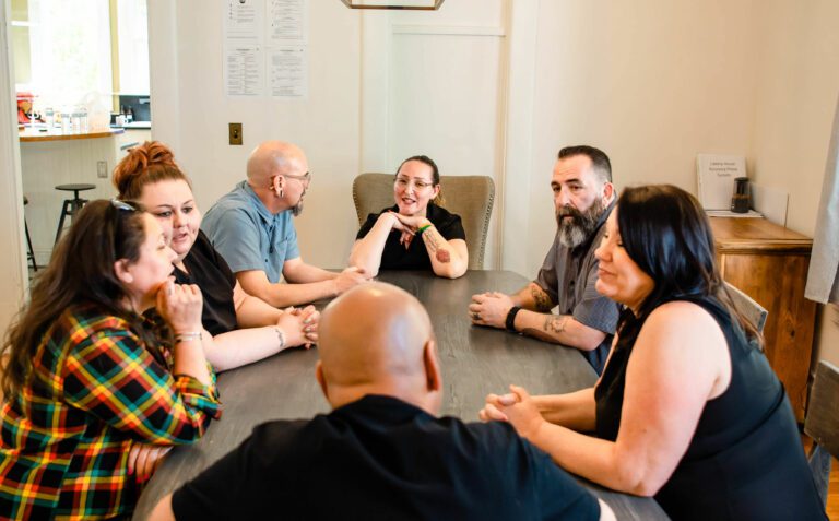 A group of people engaged in a lively conversation around a dining table in a cozy room at an addiction recovery center.