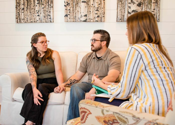 A couple sitting on a white couch conversed earnestly with a woman across from them, who appeared to be taking notes during a consultation or therapy session.